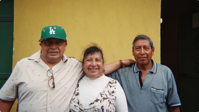 Three people smiling in front of a yellow wall, one wearing a green LA cap.